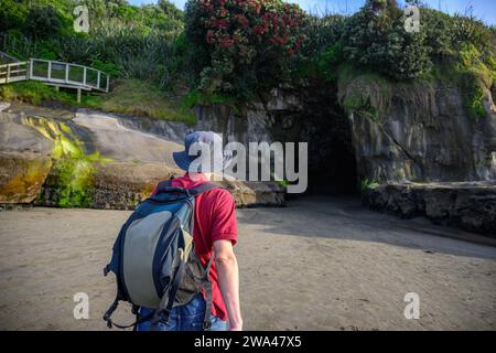 Man walking towards the entrance of Muriwai cave. Pohutukawa trees in bloom. Muriwai Beach in summer. Auckland. Stock Photo