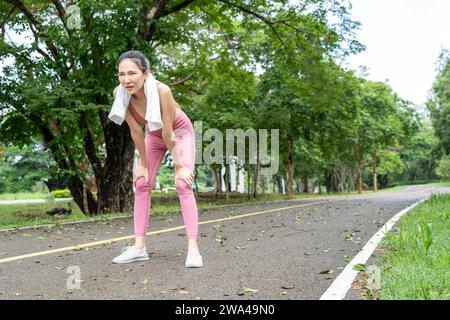 Tired young Asian woman in fitness clothes stops and leans down to take a break from her morning run at a running track of a local park Stock Photo