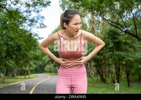 Young atractive Asian woman in fitness clothes with stomach pain putting her hands over her stomach while standing at a running track of a local park Stock Photo