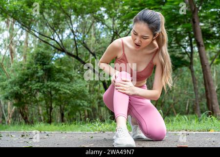 Young atractive Asian woman in fitness clothes with knee pain putting her hands over her troubled right knee while sitting down at a running track of Stock Photo