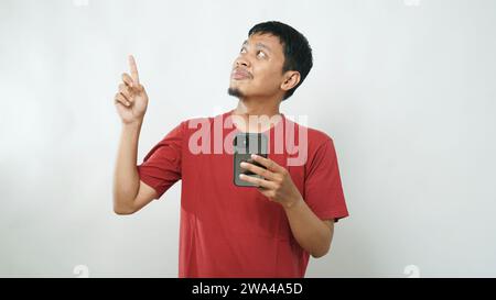 Young Asian man using smartphone over white background with a big smile on face, pointing with hand finger to the side looking at the camera. studio s Stock Photo
