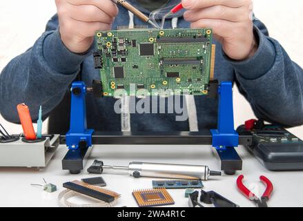 Close up of a technician's hands in a workshop. Repairer is soldering circuit board of electronic device on table with tweezers. Stock Photo