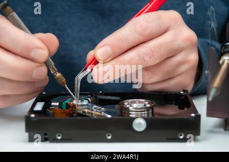 Close-up of a technician's hands in a workshop. The technician is using tweezers and a screwdriver to repair and maintain a hard drive. Stock Photo