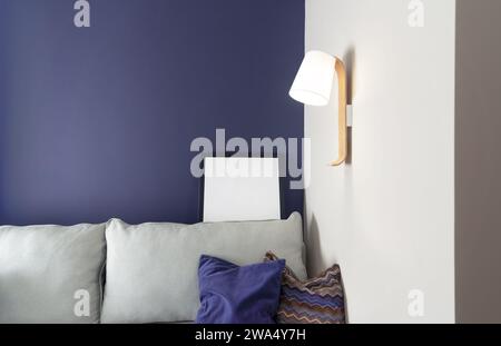 Living room interioir in a minimalist style with soft blue walls, sofa with fabric cushions, lamp on the wall and mockup frame. Stock Photo