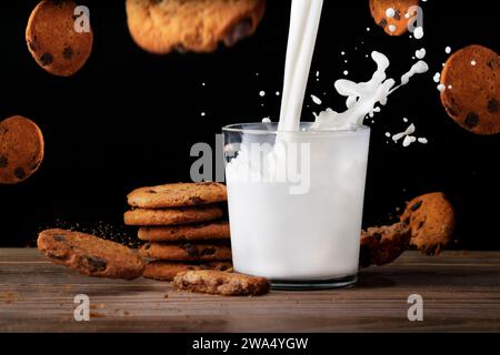 Glass of milk with tasty cookies. Milk is poured into a cup with splashes. Cookies flying in the air and fall on the table with crumbs. Black backgrou Stock Photo