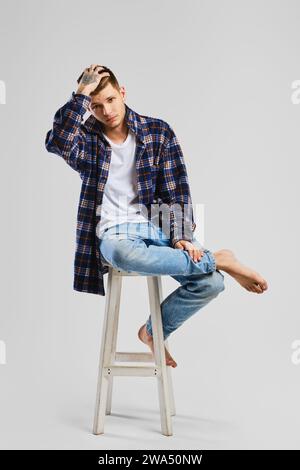 Full length studio portrait of young cocky man in shirt and jeans sitting on tall wooden chair Stock Photo