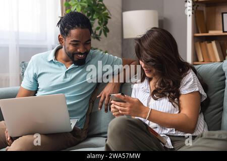 Cheerful millennial mixed couple spending time with laptop and book at home, happy young interracial lovers sitting on sofa with computer, freelance w Stock Photo
