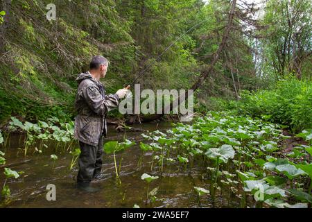 fisherman with a spin fishing rod in his hands stands in the middle of a narrow forest river Stock Photo