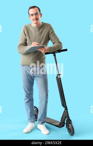 Young man with kick scooter writing in clipboard on blue background Stock Photo