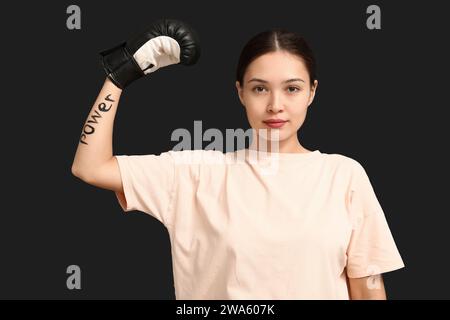 Young woman in boxing glove with word POWER written on her arm against black background. Feminism concept Stock Photo