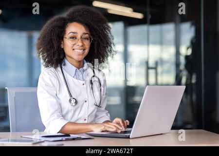 Portrait of a young African American female doctor in a white coat sitting at a table and working at a laptop, looking at the camera smiling. Stock Photo