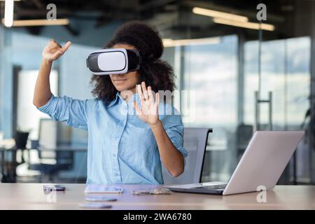 Young African American woman sitting in the office at the table with a laptop and talking online through a virtual mask, gesturing with her hands. Stock Photo