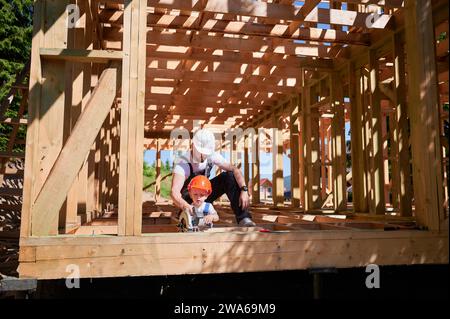 Father with toddler son building wooden frame house. Male builder teaching his son how to cut board using electric saw on construction site, wearing helmets and overalls. Carpentry and family concept. Stock Photo