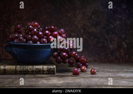 Fresh red grape in a blue bowl with a vintage book on wooden board table and background - dark and moody Stock Photo