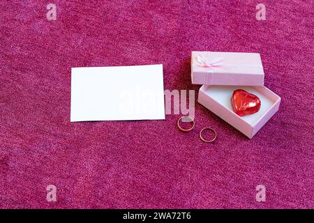 open gift box with a heart-shaped chocolate inside and engagement rings on a dark pink background Stock Photo