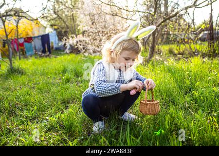 Small girl wearing Bunny ears find and pick up egg on Easter egg hunt in garden Stock Photo