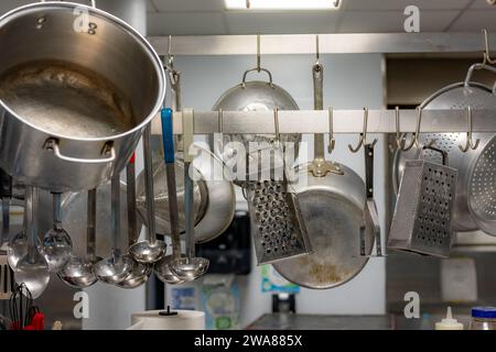 Stainless steel pots and pans and other cooking utensils hanging in a commercial industrial kitchen. Stock Photo