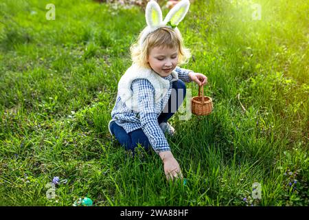 Easter tradition. A girl with a basket collects colorful Easter eggs in the grass. Child wearing bunny ears. Stock Photo