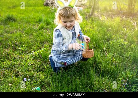 girl wearing bunny ears playing egg hunt on Easter. A child celebrates Easter. Girl sitting on grass with a basket full of colorful eggs. Easter egg h Stock Photo