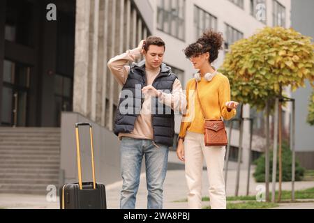 Being late. Worried man and woman with suitcase looking at watch outdoors Stock Photo