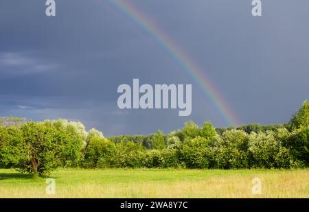 Real rainbow in dark sky. Countryside landscape with fields and trees. Stock Photo