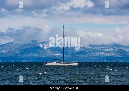 A sailboat anchored on lake Tahoe after a late spring snow storm, with cloudy skies and fresh new snow on the mountains in the background. Stock Photo