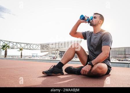Weary sports man resting and drinking water after endurance workout sitting on the floor. Exhauted male recovering and having a routine break taking a protein supplement at cardio wellness training. High quality photo Stock Photo