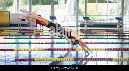 Professional female swimmer preparing and jumping off the starting block into the pool. Competitive swimmers workout concept. Stock Photo