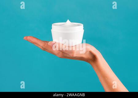 Anonymous young slim female hand showing white jar with body cream in palm with manicured fingers in light against turquoise background Stock Photo