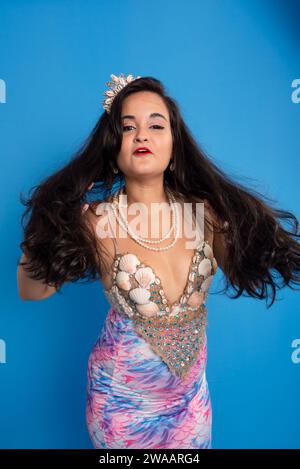 Beautiful woman, wearing a tiara and seashell bracelets, against a blue background, touching her black hair. Yemanja worshiper. Stock Photo