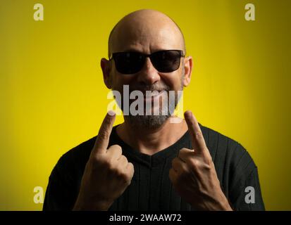 Portrait of bald, bearded mature man wearing sunglasses making sign with his hands. Isolated on yellow background. Stock Photo