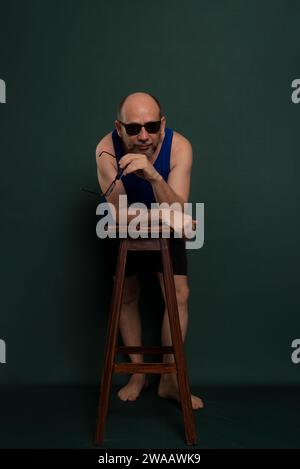 Bearded, bald white man wearing sunglasses leaning on wooden stool posing for photo. Isolated on dark green background. Stock Photo