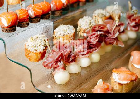 A luxurious array of gourmet canapes, including melon with prosciutto and smoked salmon on pumpernickel, presented on a chic glass serving tray. Stock Photo