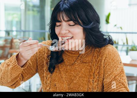 young Latin Venezuelan woman with long black hair dressed in brown sweater, sitting in cafeteria smiling happily eating slice of chocolate cake, food Stock Photo