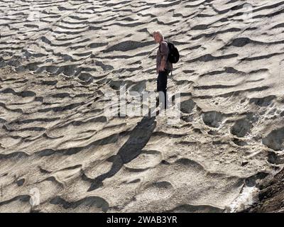 lone female hiker in Summer sunshine casting long shadow across scalloped snow standing in deep foot-holes on ice sheet in foothills of Italian Alps Stock Photo