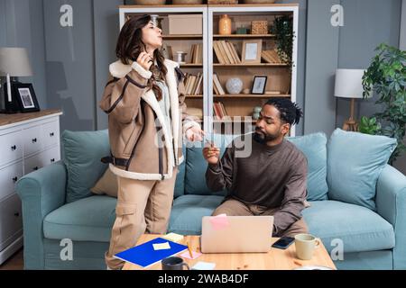 Young woman asking for credit card from her husband to go to shopping while he working from home on laptop. Freelance male unwillingly giving his bank Stock Photo
