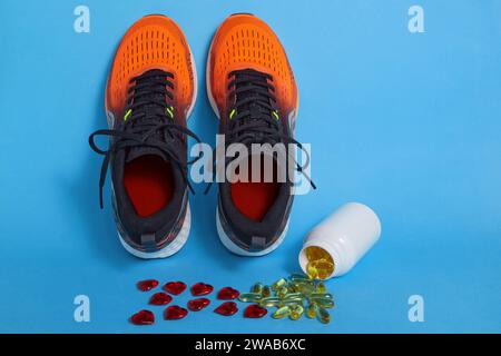 Scattered yellow vitamin D capsules from a jar and red decorative hearts and orange running sneakers on a blue background. Stock Photo