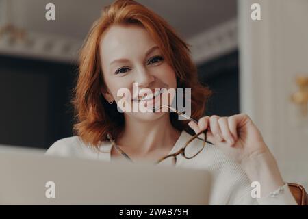 Redheaded woman in white, holding glasses, beams at her laptop, exuding warmth and confidence in her gaze Stock Photo