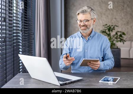 Senior tired man sits in the office in front of a laptop, talks, studies and works remotely online, holds and writes in a notebook. Stock Photo