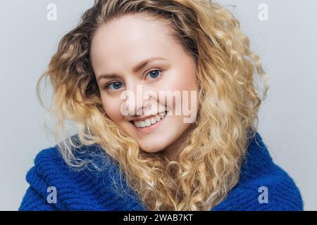 Cheerful young woman with vibrant curly blonde hair, beaming smile, wrapped in a cozy blue knit sweater. Stock Photo