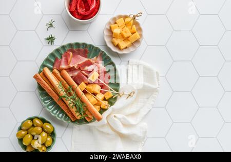 Slices of prosciutto or jamon. Antipasto. Delicious grissini sticks with prosciutto, cheese, rosemary, olives on green plate on white concrete backgro Stock Photo