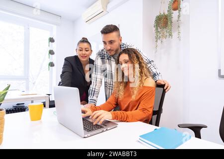A multi-ethnic team of three collaborates around a laptop in a bright office adorned with green plants Stock Photo