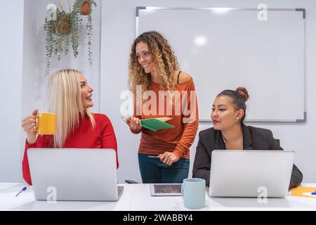 Smiling businesswomen discuss over laptops with coffee mugs, in a well-lit office space.  Stock Photo