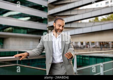 Confident businessman in grey suit using smartphone while leaning on railing outdoors Stock Photo