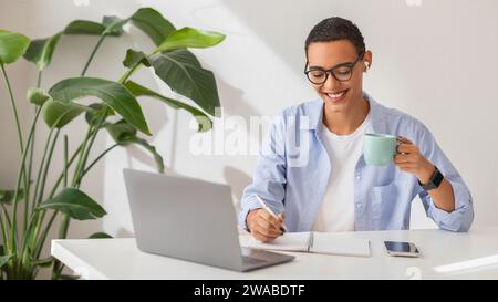 Joyful young professional taking notes while holding a coffee mug, wearing a casual blue shirt Stock Photo