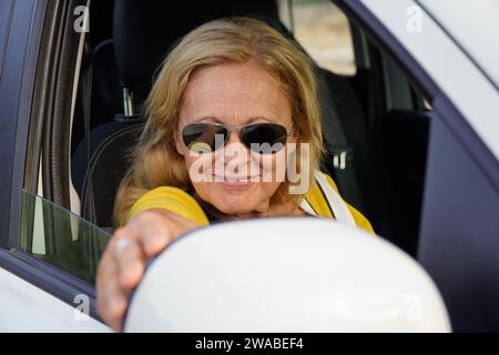 Mature woman with sunglasses and summer clothes adjusting the car rearview mirror Stock Photo