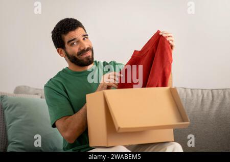 Smiling young man in a green shirt skeptically examines a red sweater just unpacked Stock Photo