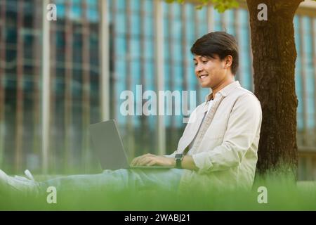 Asian guy freelancer sitting under tree, working on laptop Stock Photo
