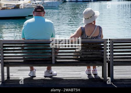 Barcelona, Spain - May 26, 2022: Senior couple contemplate sitting on a bench next to the marina, equipped with sneakers, cap and hat. Stock Photo