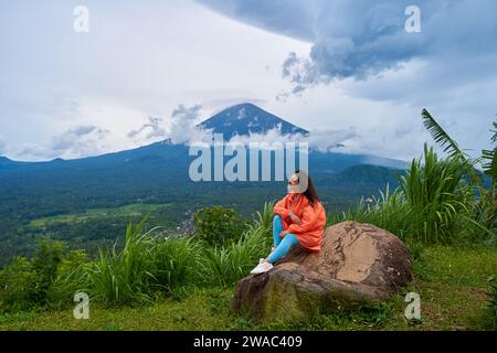A young woman sits on a large rock in a viewpoint and enjoys the view of the sacred Mount Agung volcano hidden by clouds on a rainy day on the island Stock Photo